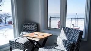 Una habitación con dos sillones estampados y una pequeña mesa, un ventanal con vistas al mar y una estructura metálica, además de una planta en maceta sobre el alféizar.