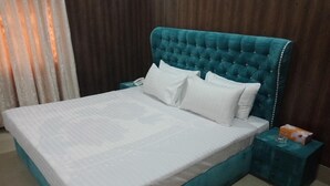 A neatly made bed with a teal headboard and white bedding, a small teal nightstand with a tissue box, and a wooden wall with a curtain partially visible. - Hotel Royal Defence (Lahore)