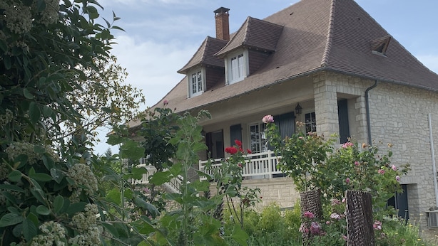 A stone house with a tiled roof, a porch with a railing, and a garden with various plants and flowers.