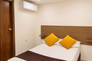 A single bed with a wooden headboard, two yellow pillows, a brown throw blanket, and a wall-mounted air conditioning unit.