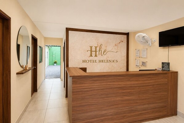 A hotel reception desk with a wooden finish, a wall-mounted TV, and a reception area with a green outdoor view.