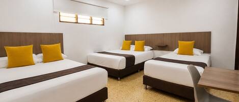 A hotel room with three beds, each with white bedding and yellow pillows, a wooden headboard, a small table, and a chair.