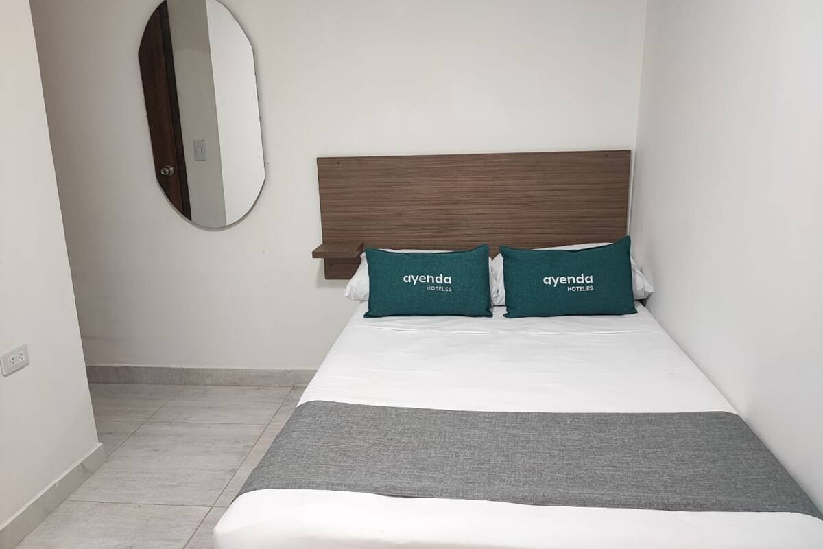 A hotel room with a bed, two pillows with 'ayenda HOTELES' text, a wooden headboard, a round mirror, and a grey blanket.