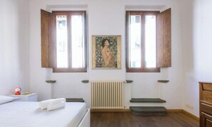 Um quarto com uma cama, duas janelas, um radiador e um quadro na parede.