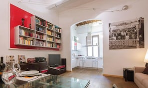 Sala de estar com estante, televisão, mesa de vidro e cozinha visível por uma abóbada.