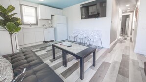 Ein modernes Wohnzimmer mit einem schwarzen Sofa, einem Couchtisch und einem Esstisch. Es gibt eine Küchenzeile mit weißen Schränken und einem Kühlschrank. Im Eckbereich steht eine große Pflanze, und im Essbereich befinden sich weiße Stühle.