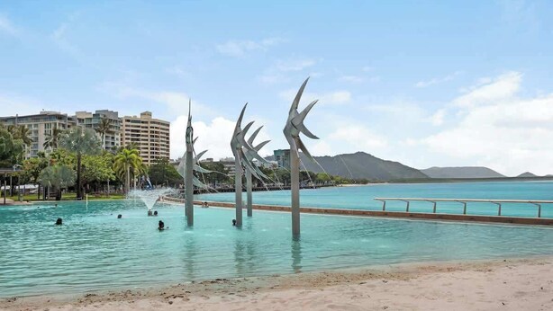 A pool area with clear water, a sandy beach, and a backdrop of buildings and mountains.