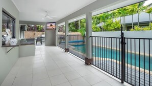 A spacious indoor area with a pool, a ceiling fan, and a view of a wooden fence and greenery. - Tropical Pool Sanctuary in Kewarra Beach (Kewarra Beach)