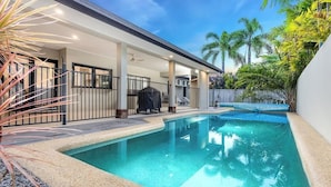 A swimming pool with a covered patio area, surrounded by tropical plants and a clear sky. - Tropical Pool Sanctuary in Kewarra Beach (Kewarra Beach)