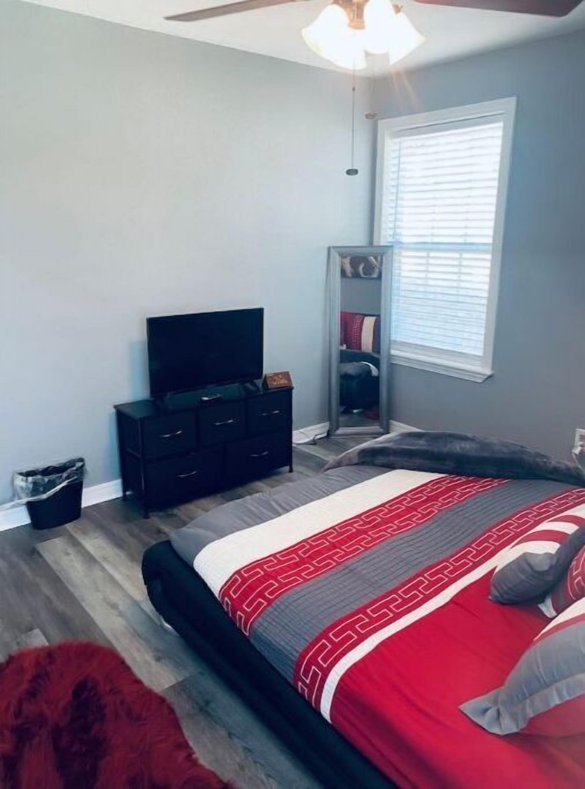 A bedroom with a bed, a dresser, a television, a window with blinds, and a ceiling fan.
