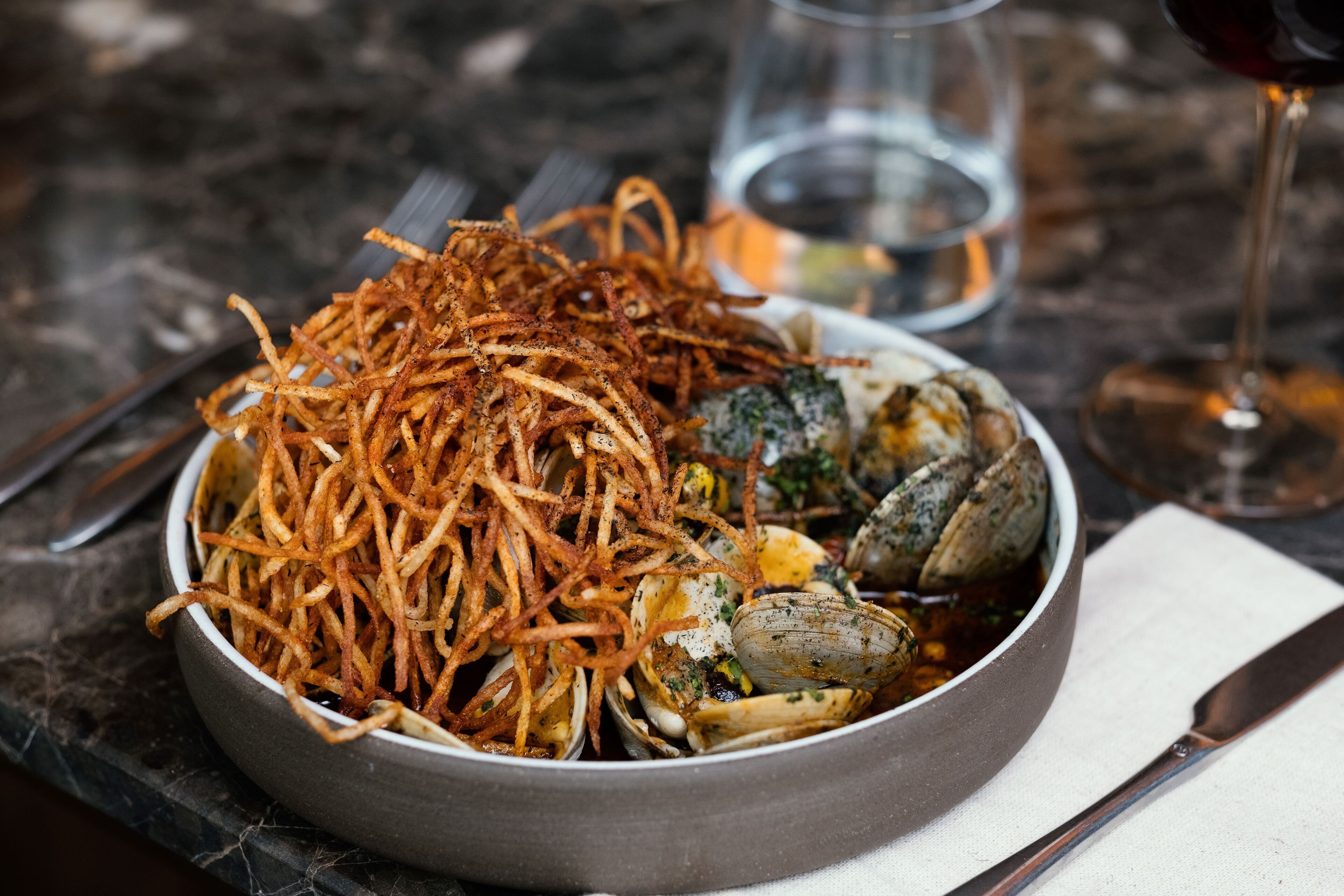 A bowl of food with fried potatoes and clams, accompanied by a glass of red wine and a fork.
