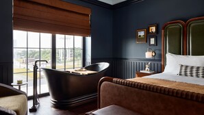 A room with a large window, a bathtub, a bed with a headboard, and a small table with a lamp. - The Manchester  (Lexington)