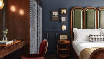 Habitación de hotel con un escritorio de madera, una silla roja, una cama con almohadas blancas y estampadas, una mesita de noche con una lámpara y cuadros en la pared.