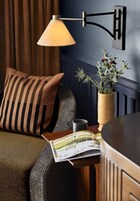 Un rincón de estar acogedor con un cojín a rayas, una mesa con una revista, un florero con flores y una lámpara de pared.