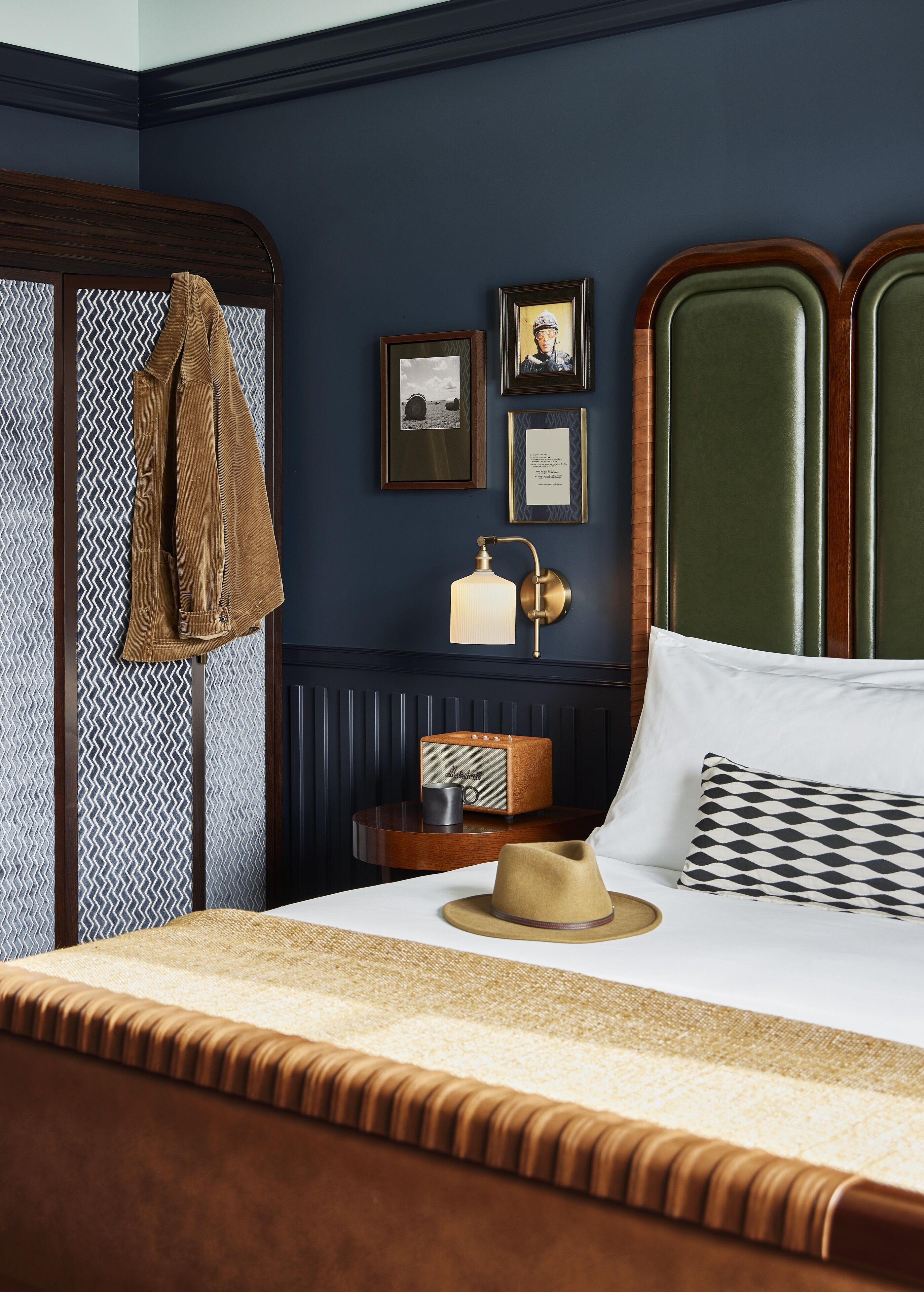 A neatly arranged bedroom with a bed, a side table with a lamp, a radio, and a hat on the bed.