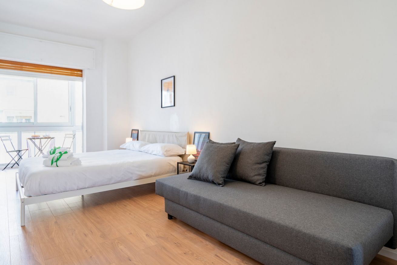 Ein modernes Hotelzimmer mit einem Bett, einem grauen Sofa, einem kleinen Tisch mit einer Lampe und einem Fenster mit Jalousien.