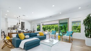Sala de estar moderna com sofá azul, mesa de centro de vidro e vista para uma piscina e vegetação lá fora.