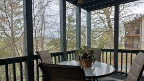 A covered outdoor seating area with a round table and four chairs, a wicker basket with plants, and a view of trees and a building in the background. - Pools~Screened-In Deck~Dog-Friendly~2 King Beds (Branson)