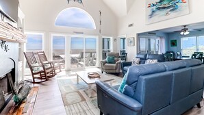 A spacious living area with a high ceiling, a large window, and a view of the ocean. There is a blue sofa, a wooden rocking chair, and a dining table with chairs. - El Mar! Beachfront - Every room is oceanfront with it's own porch & bathroom access! (Folly Beach)