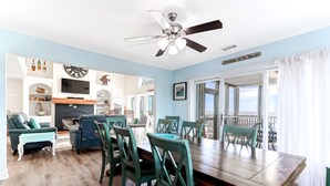 A spacious dining area with a wooden table and teal chairs, a ceiling fan, and a view of the outdoors through large windows. - El Mar! Beachfront - Every room is oceanfront with it's own porch & bathroom access! (Folly Beach)
