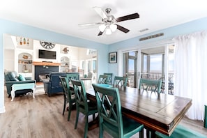 Un vaste espace dédié à la salle à manger, avec une table en bois et des chaises de couleur turquoise, un ventilateur de plafond et une vue sur l’extérieur grâce à de grandes fenêtres.