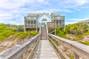 Une maison à deux étages située en bord de mer, avec une terrasse en bois menant à l’entrée.