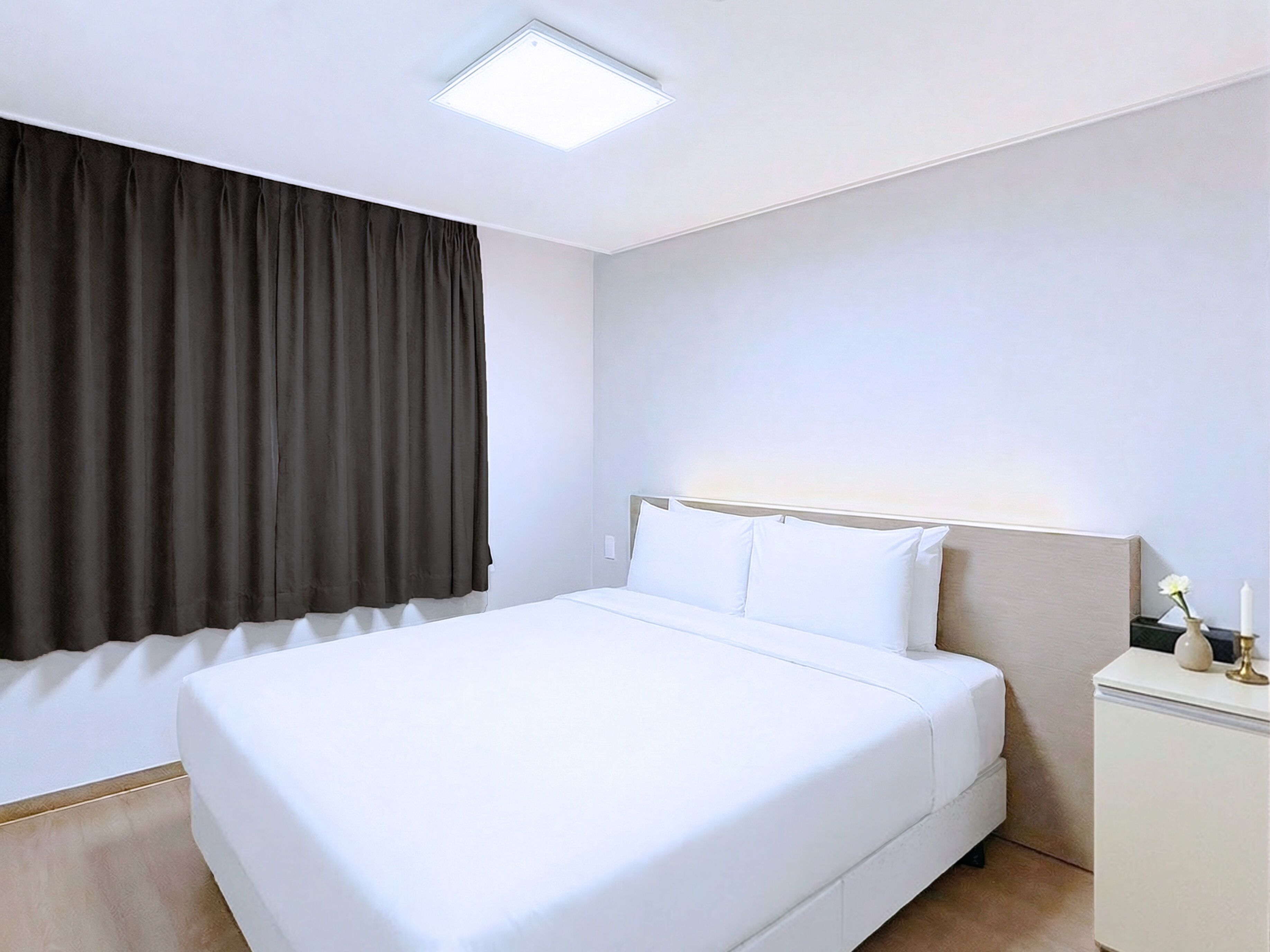 Uma cama bem arrumada num quarto de hotel, com uma janela grande coberta por cortinas escuras, uma mesa de cabeceira com abajur e uma luminária no teto.