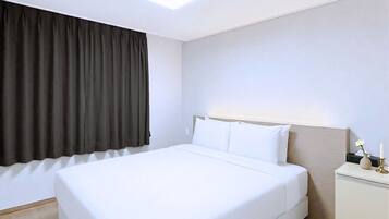 Uma cama bem arrumada num quarto de hotel, com uma janela grande coberta por cortinas escuras, uma mesa de cabeceira com abajur e uma luminária no teto.