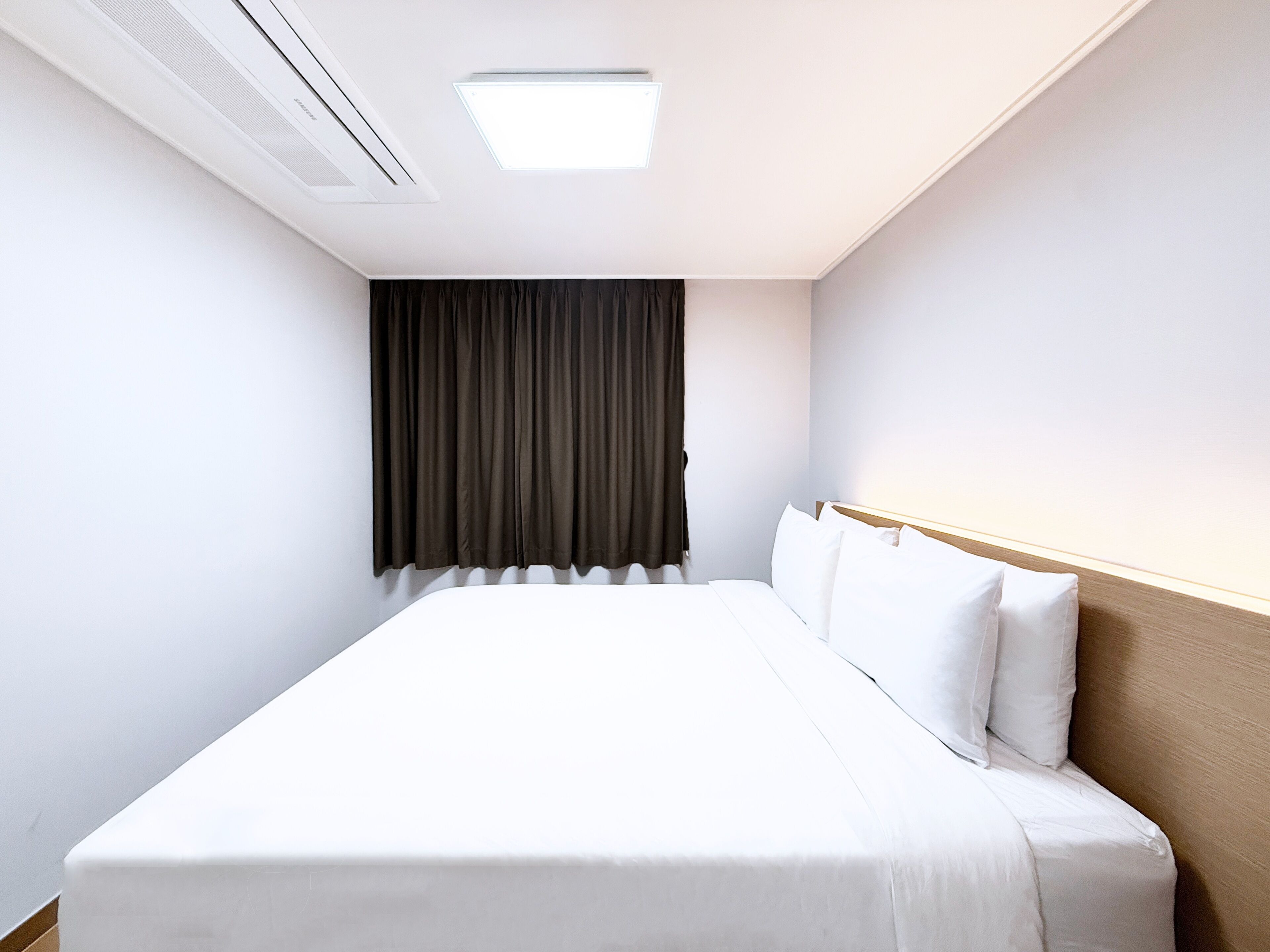 Habitación de hotel con cama, cabecera, ventana con cortinas e iluminación en el techo.