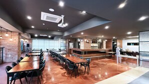 Una amplia sala de conferencias con mesas y sillas largas, una zona de cocina con barra y un diseño de techo moderno.
