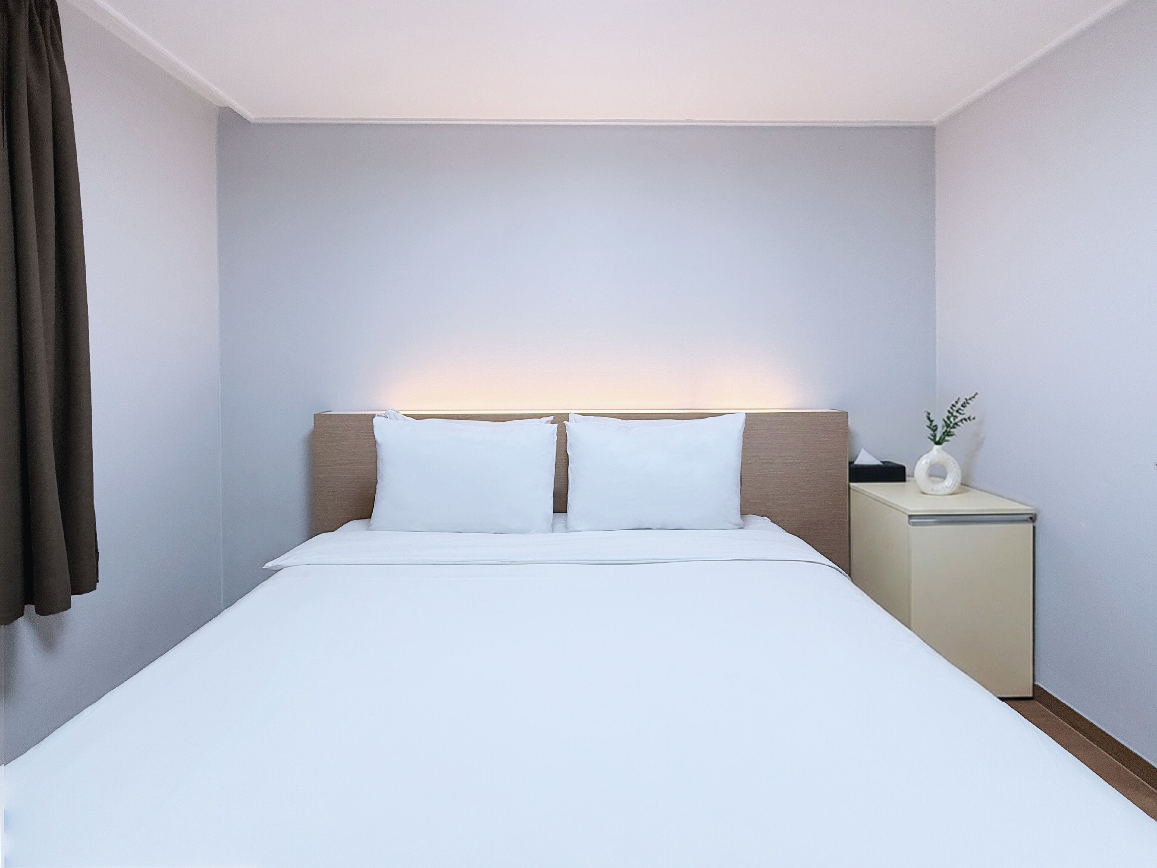 A neatly made bed with white linens and two pillows, a bedside table with a vase, and a wall-mounted light above the bed.