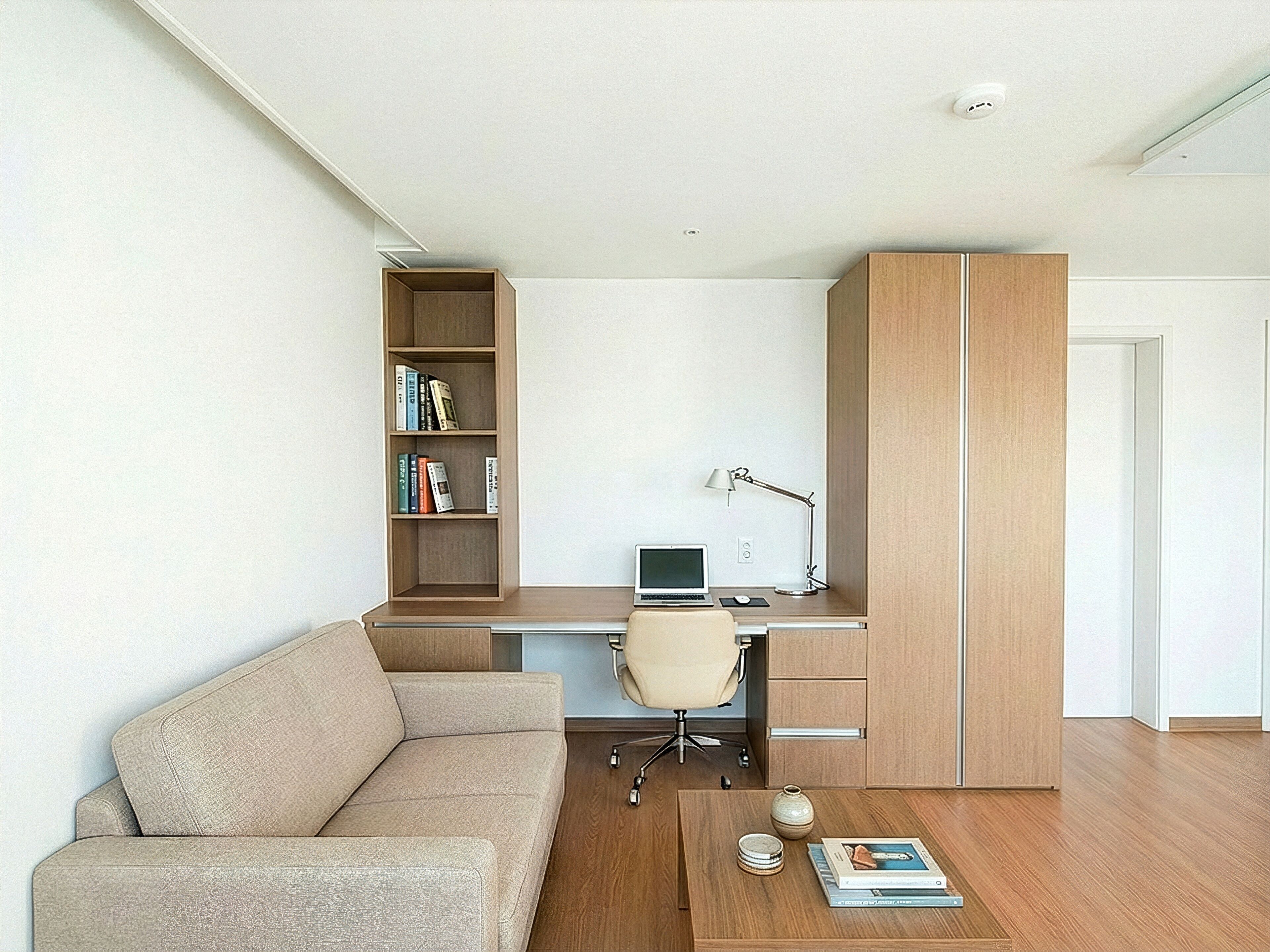 A modern, compact living space with a sofa, a desk with a laptop, a bookshelf, and a coffee table with books and a vase.