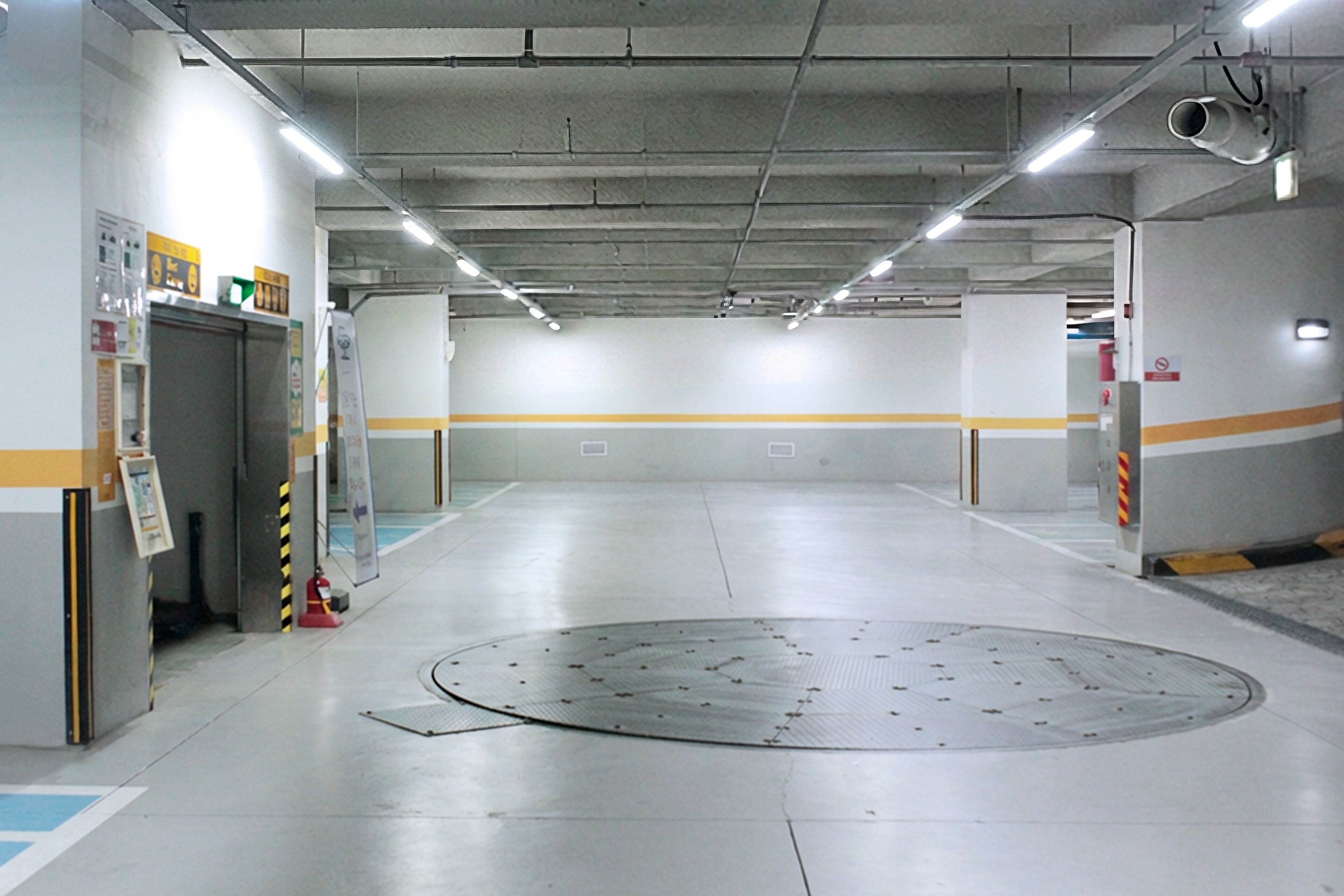 Un parking propre et bien éclairé, avec un regard d’homme circulaire au centre, des places de stationnement clairement délimitées et un extincteur sur le côté gauche.
