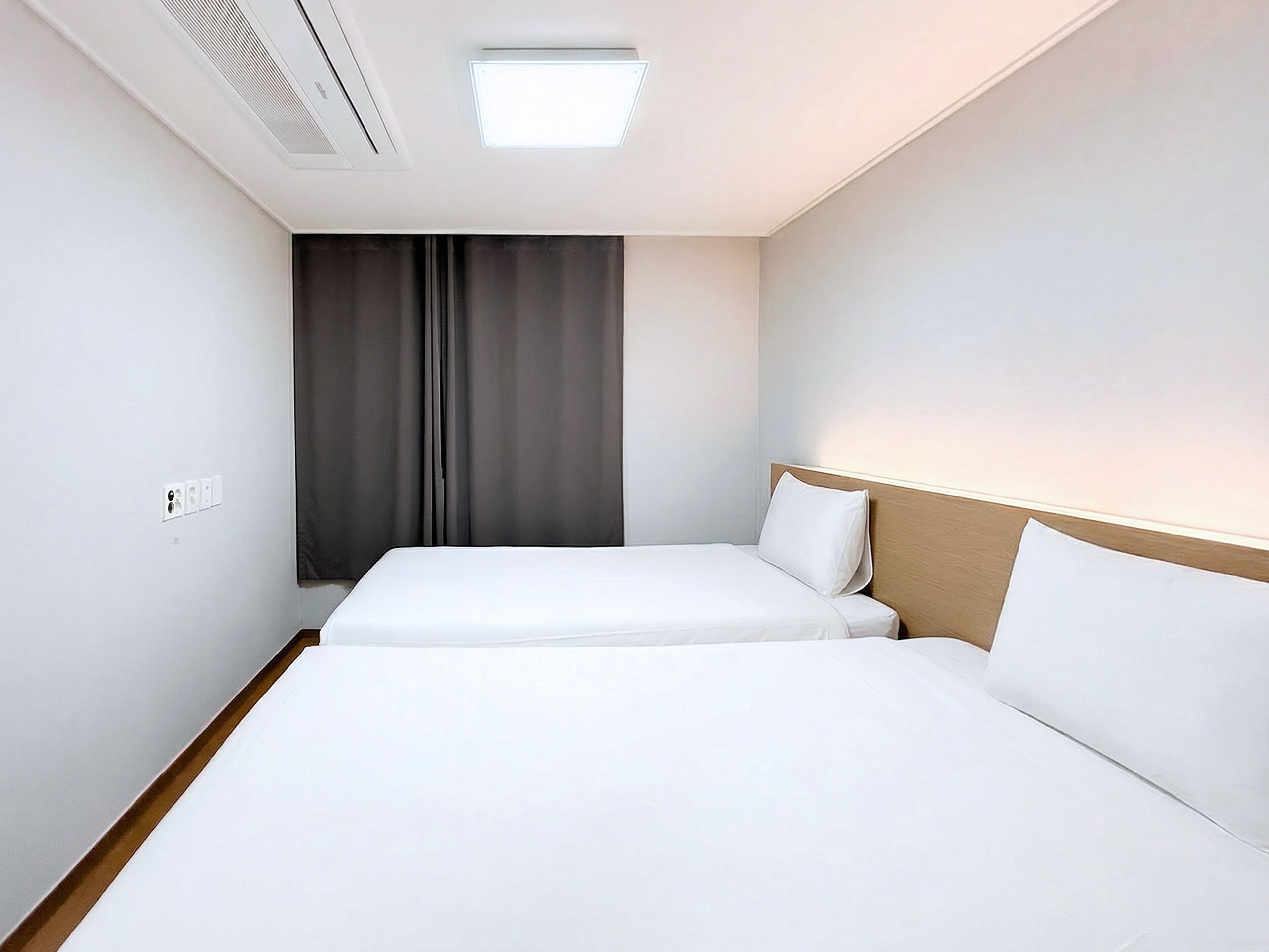 A hotel room with two single beds, a window with curtains, a ceiling light, and a wall socket.