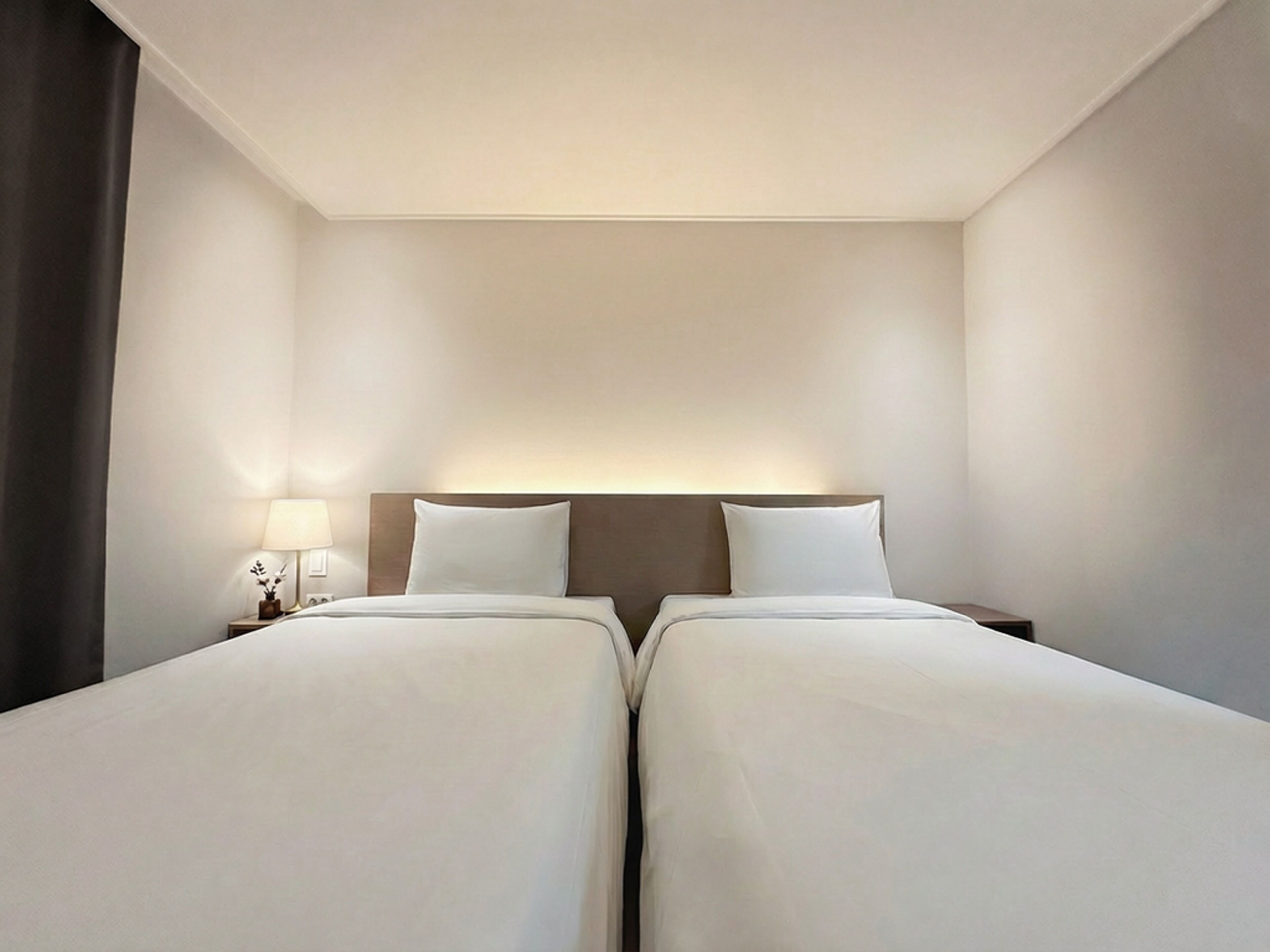 A hotel room with two beds, a nightstand with a lamp, and a dark curtain.