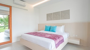 A modern bedroom with a large bed, a wooden headboard, a decorative wall piece, and a view of a garden through a glass door. - Palm Springs Bali Resorts (Tulamben)