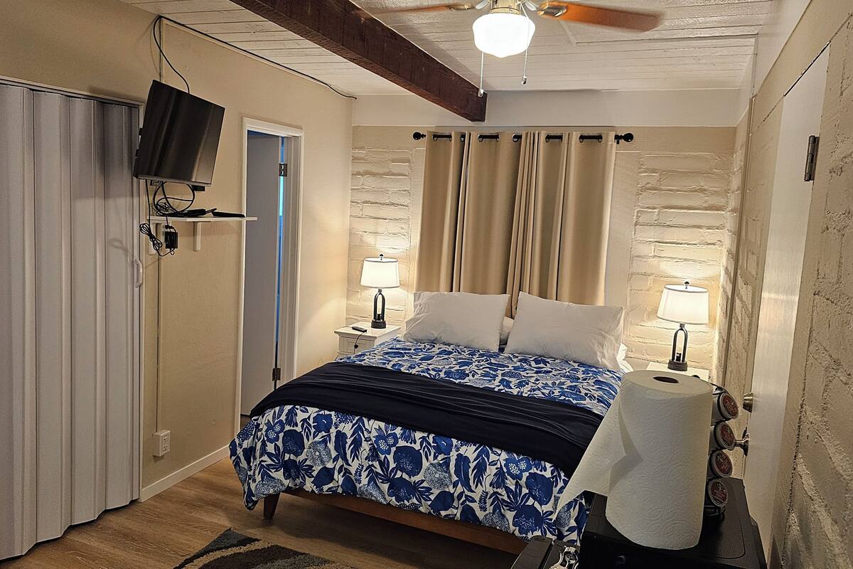 A bedroom with a bed, two bedside lamps, a ceiling fan, a TV mounted on the wall, and a door.