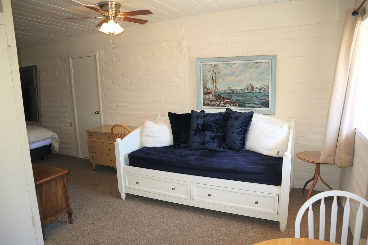 A bedroom with a white bed, a white dresser, a white chair, a white nightstand, a white sofa, a ceiling fan, a painting on the wall, and a window with curtains.