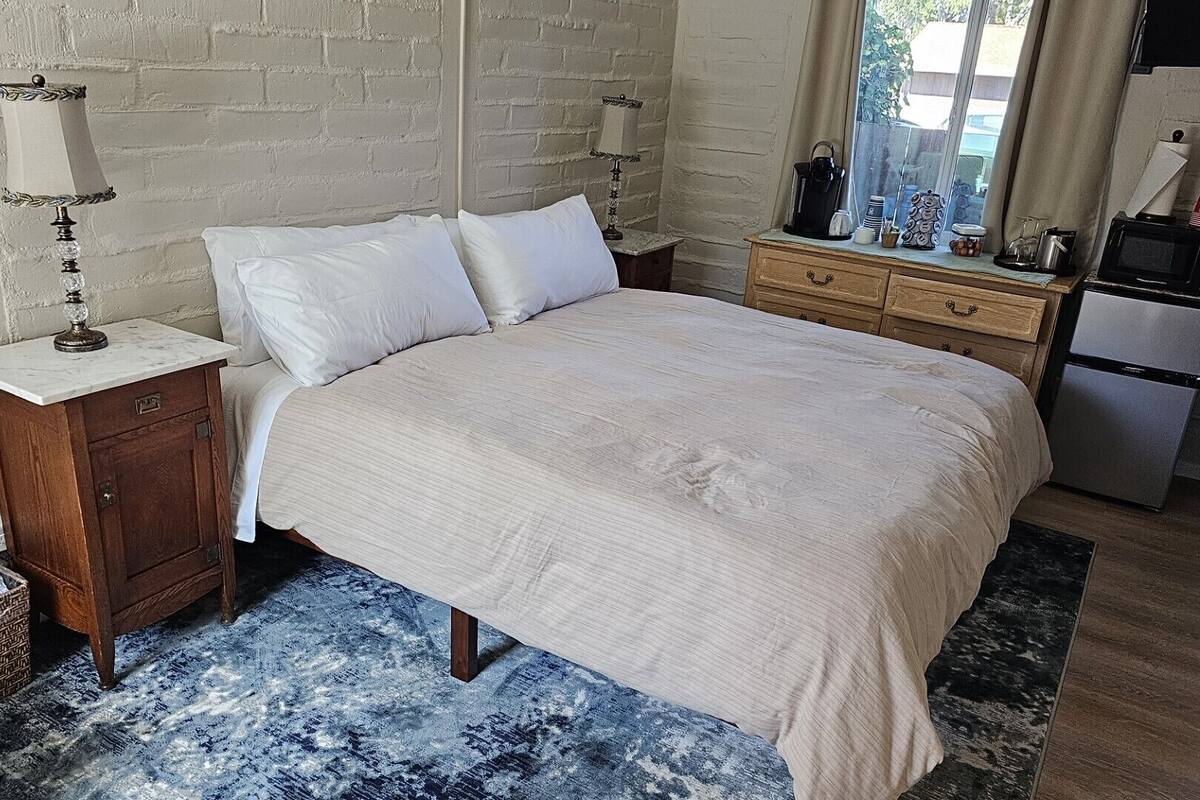 A bedroom with a bed, nightstands, a dresser, a window, and a kitchenette.
