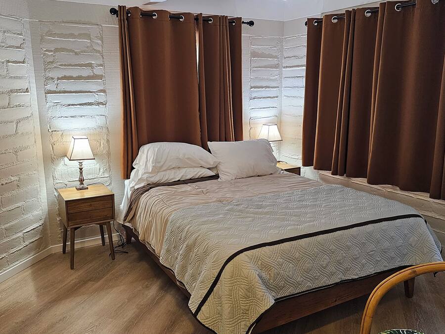 A bedroom with a bed, two bedside tables with lamps, brown curtains, and a wooden chair.