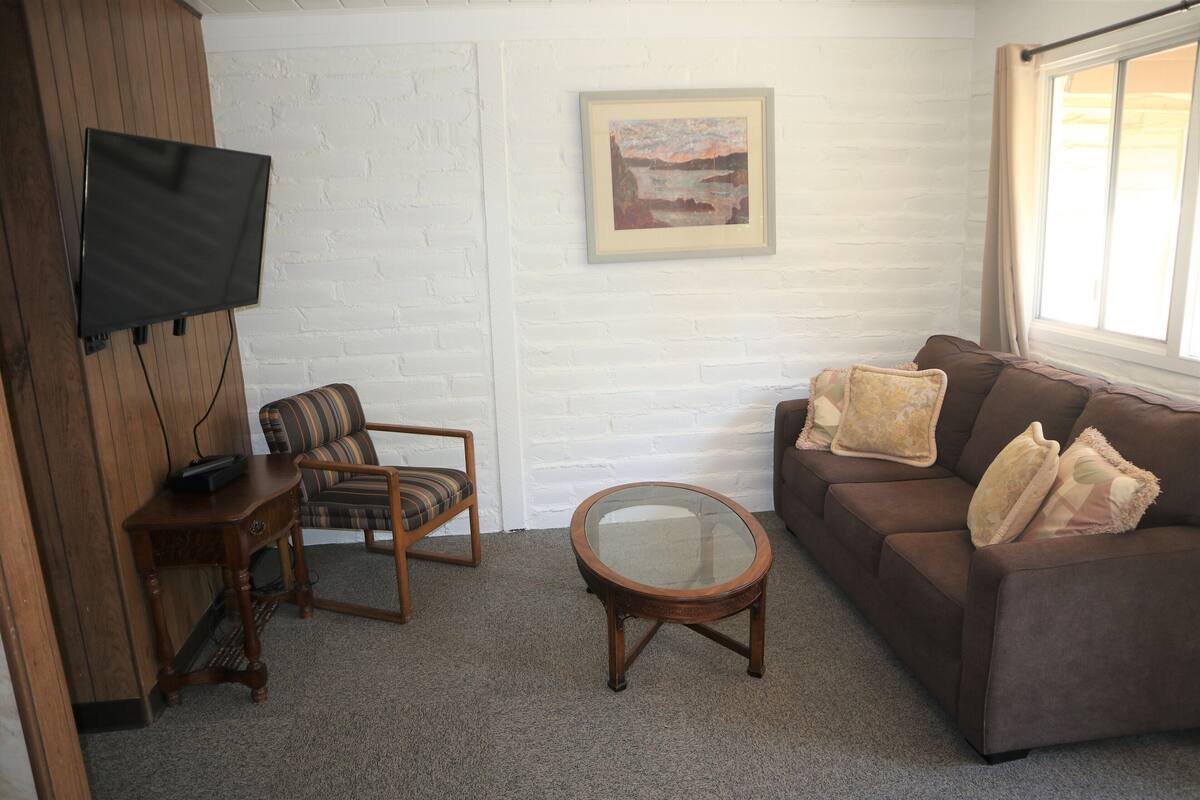 A living room with a brown sofa, a plaid armchair, a glass coffee table, a flat-screen TV mounted on a wooden wall, and a framed picture on the wall.