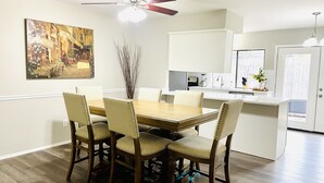 Un coin repas avec une table en bois et quatre chaises, un ventilateur de plafond, un tableau au mur et une cuisine en arrière-plan.