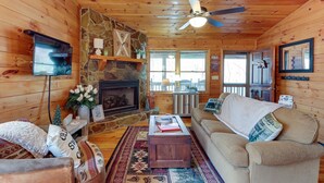 A cozy cabin interior with a stone fireplace, wooden walls, a ceiling fan, a television, and a comfortable seating area. - Private Hot Tub! ‘Cherry Beary Den’ in Blue Ridge (Blue Ridge)