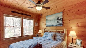 A wooden cabin bedroom with a bed, two bedside lamps, a ceiling fan, a framed picture on the wall, and a window with a view of trees. - Private Hot Tub! ‘Cherry Beary Den’ in Blue Ridge (Blue Ridge)