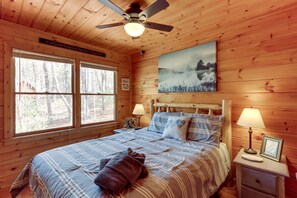 A wooden cabin bedroom with a bed, two bedside lamps, a ceiling fan, a framed picture on the wall, and a window with a view of trees.