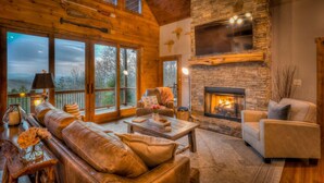 A cozy cabin interior with a stone fireplace, leather furniture, and a large window overlooking a forested landscape. - Branch Oak Manor - Million Dollar Views | Hot Tub | Fire Pit (Mineral Bluff)