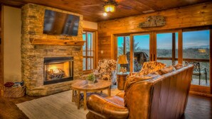 A cozy interior with a stone fireplace, leather seating, and large windows offering a view of the outdoors. - Branch Oak Manor - Million Dollar Views | Hot Tub | Fire Pit (Mineral Bluff)