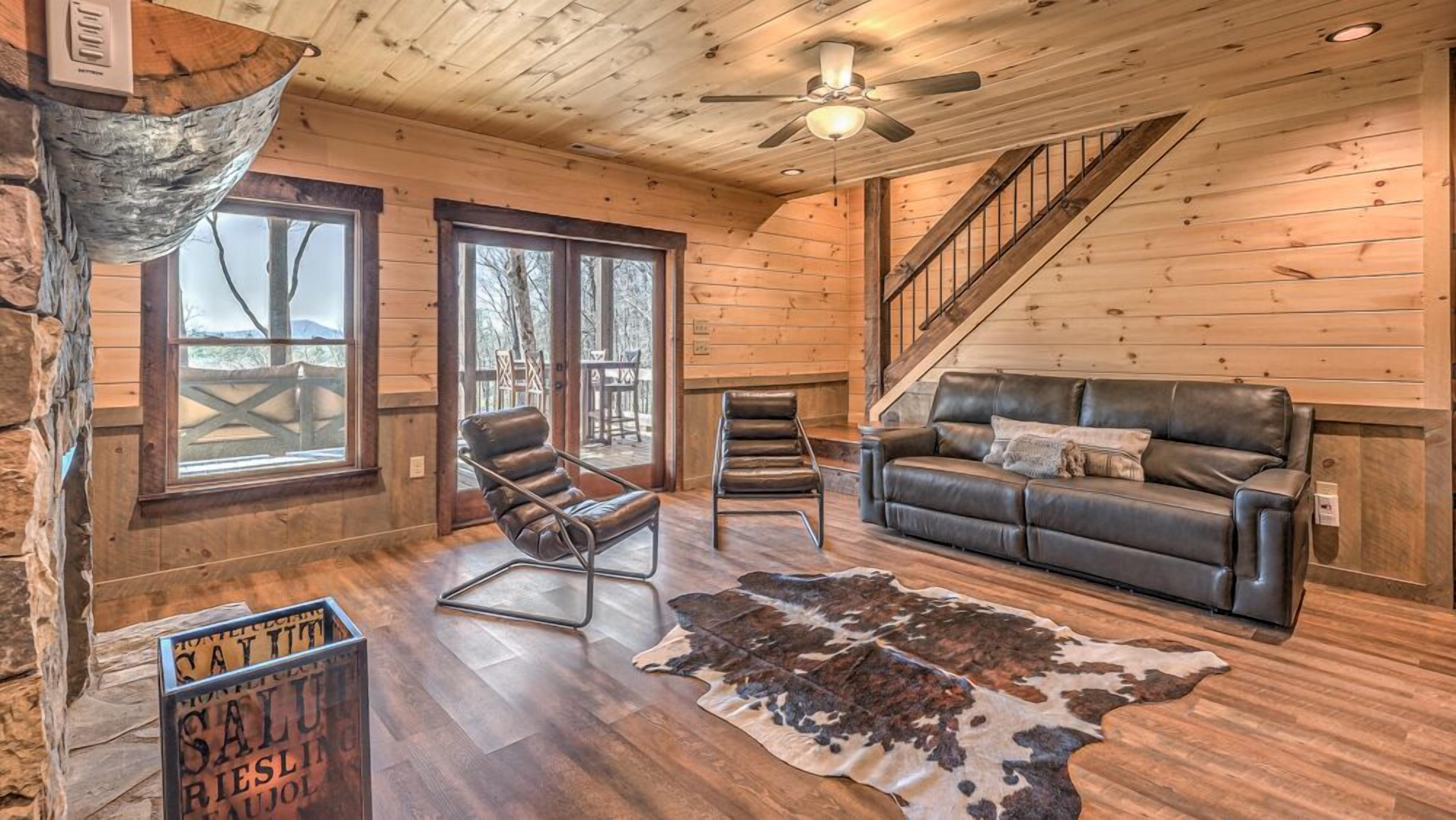L’intérieur d’une cabane en rondins, avec un canapé en cuir, deux fauteuils en cuir, un tapis en peau de vache, un ventilateur de plafond et un escalier.