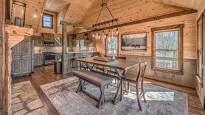 A rustic cabin interior with a wooden dining table, benches, and a stone fireplace.