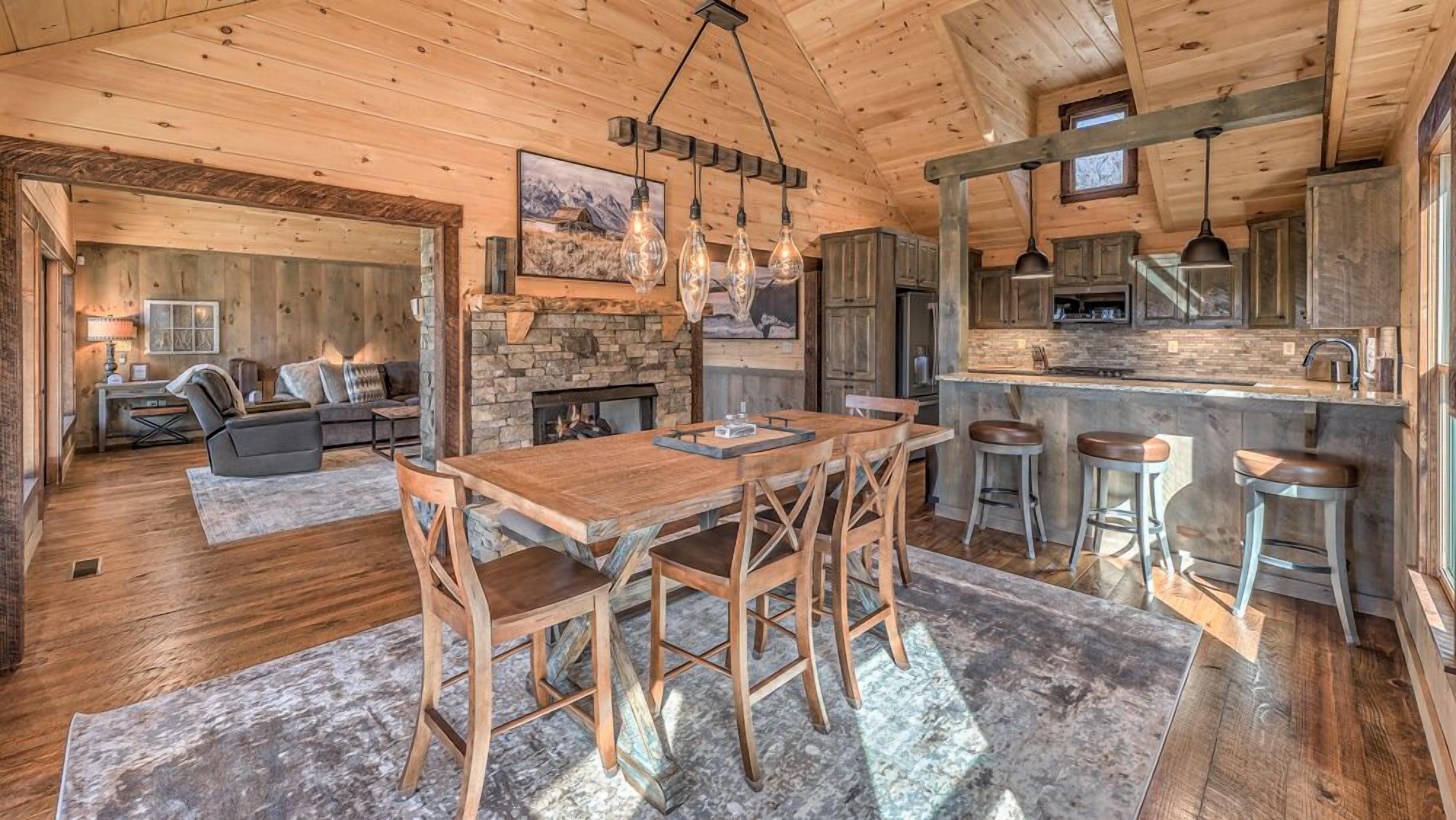 A rustic cabin interior with a wooden dining table, bar stools, a stone fireplace, and a kitchen area with a stone backsplash and wooden cabinets.